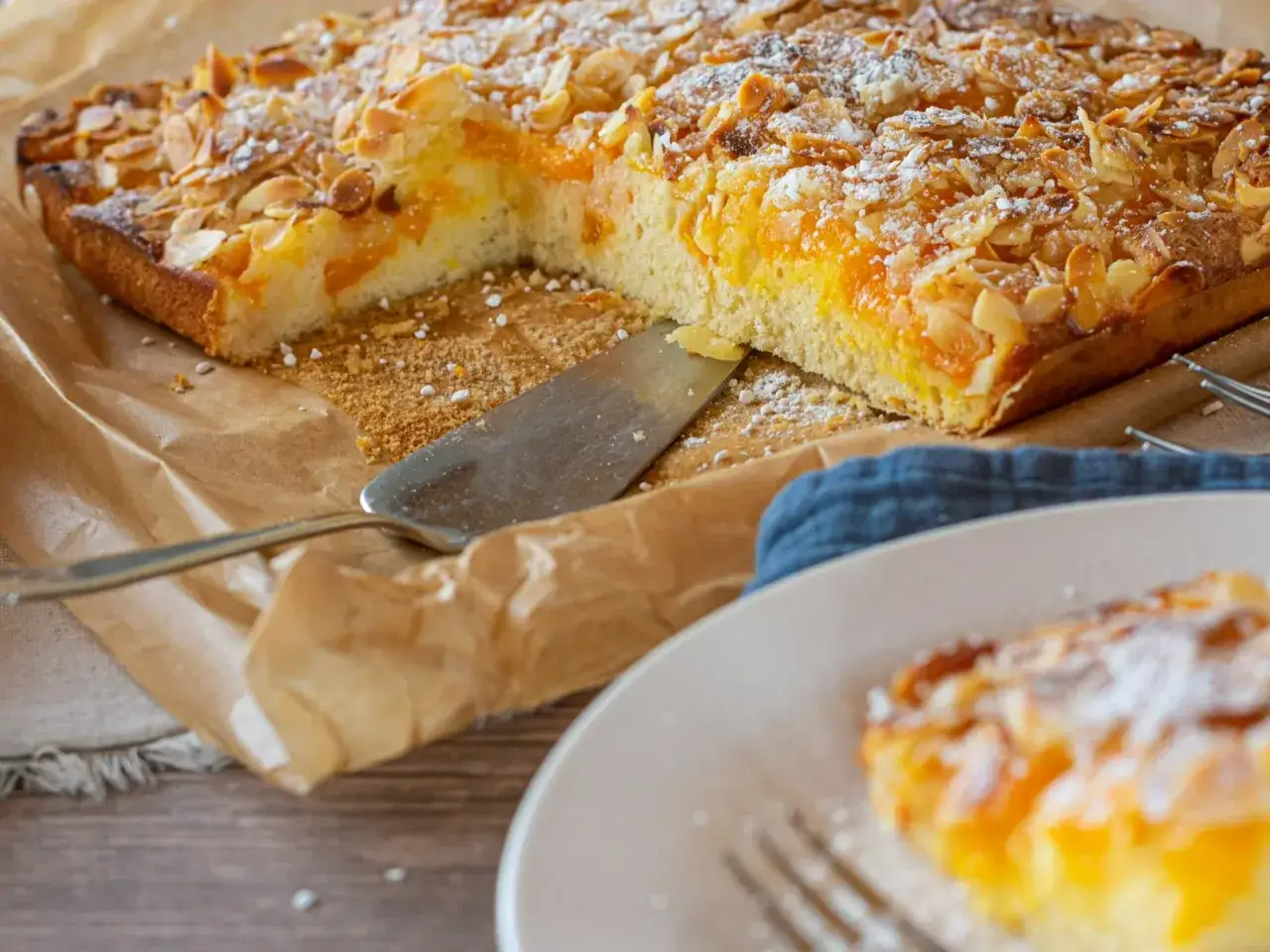 Duże ciasto z brzoskwiniami i migdałami, posypane cukrem pudrem, na papierze do pieczenia.