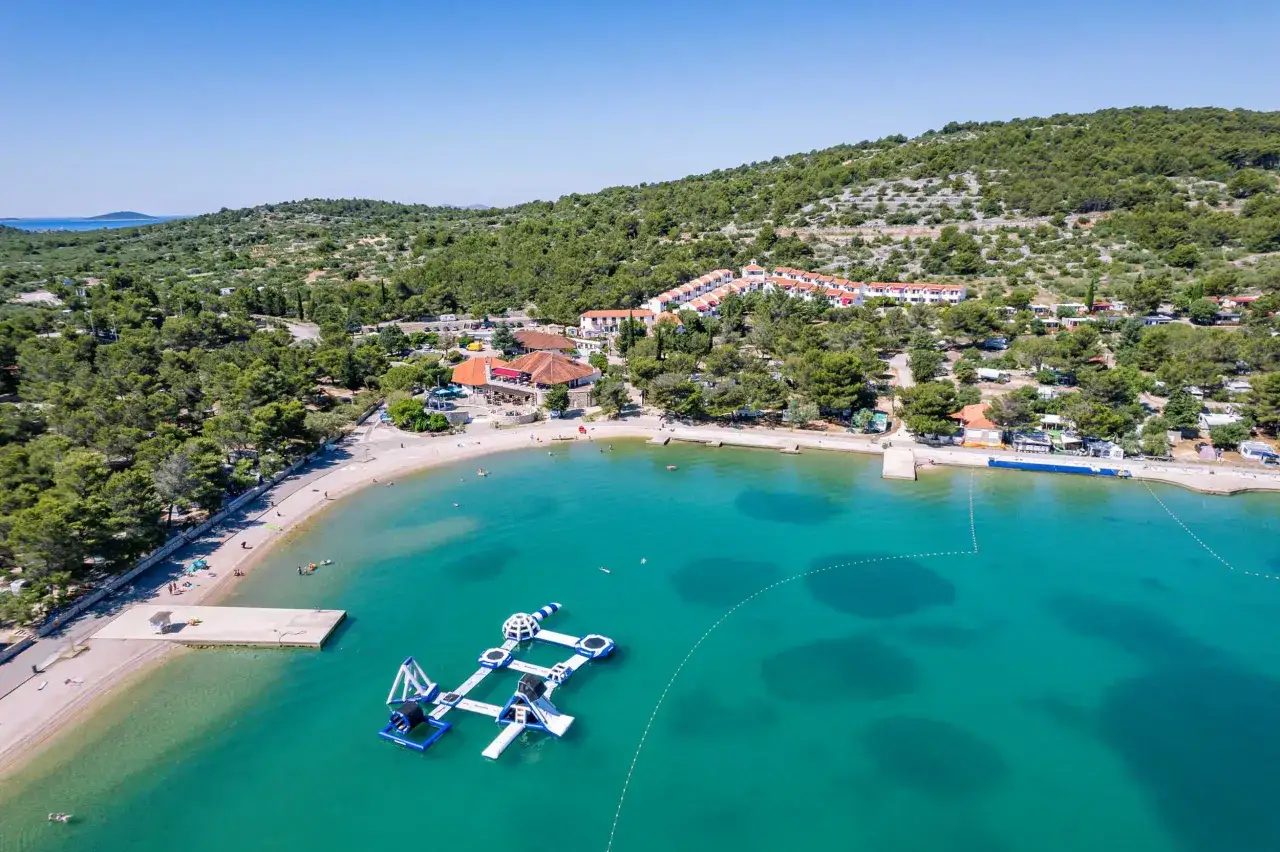 Malownicza zatoka z turkusową wodą, plażą i wodnym placem zabaw. W tle widać zabudowania i zielone wzgórza, idealne na camping Murter.