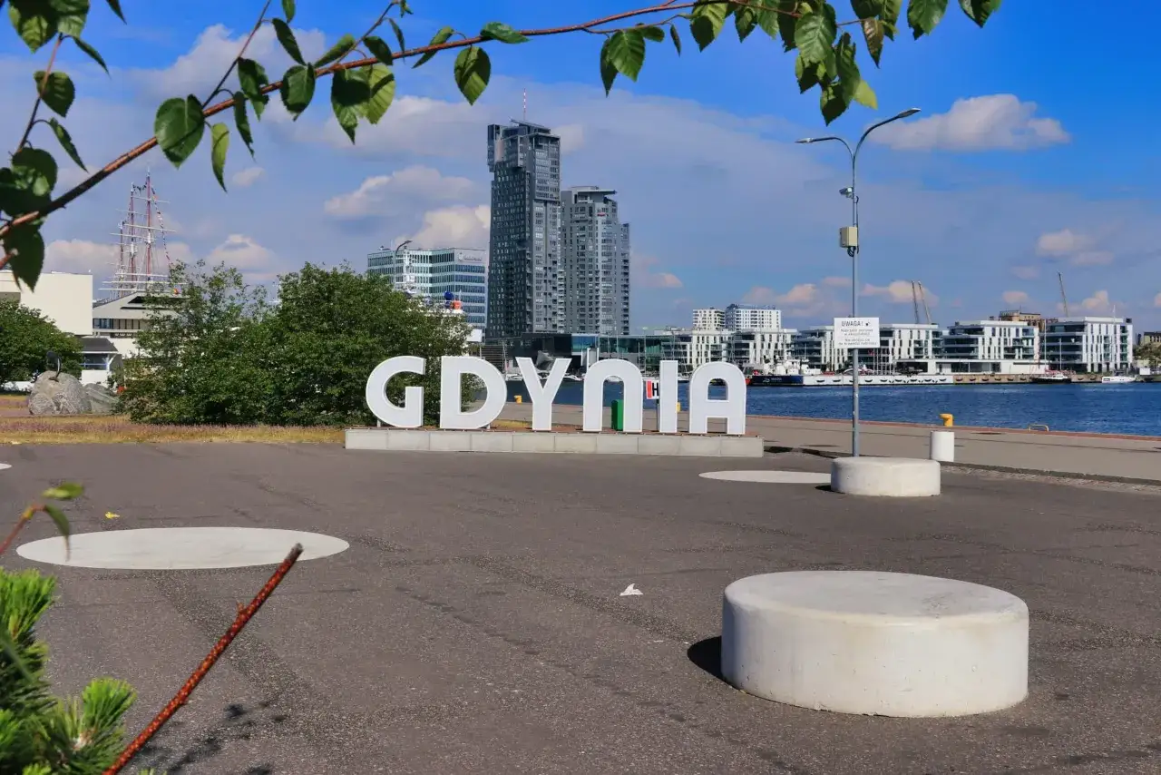 Napis GDYNIA na tle nowoczesnych budynków i żaglowca.
