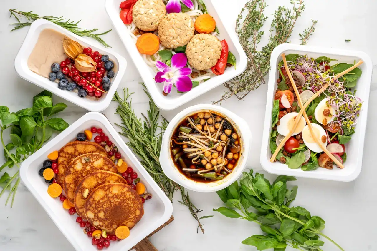 Pięć białych pojemników z różnorodnymi zdrowymi posiłkami, owocami i ziołami.