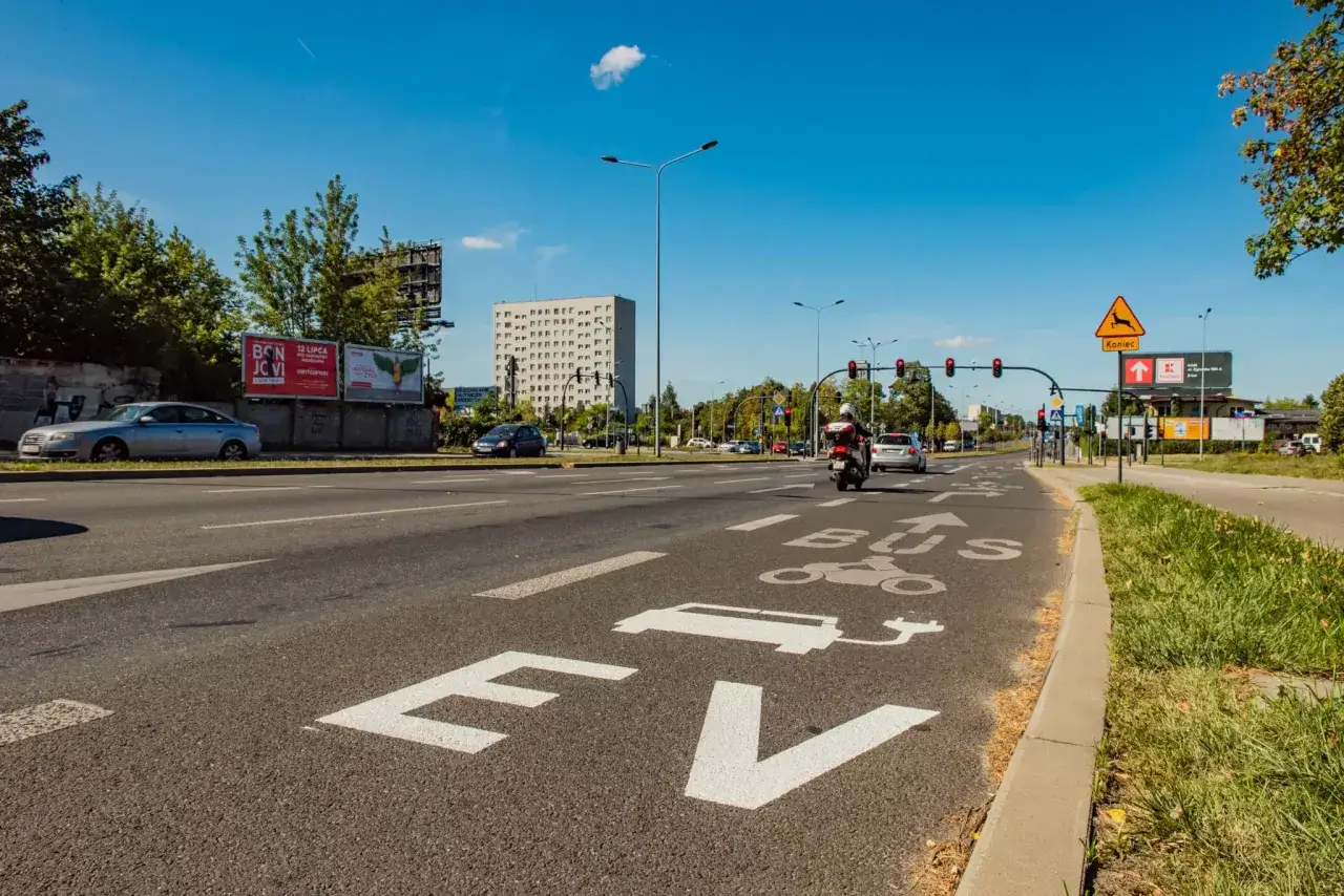Na drodze oznaczenie "EV BUS" sugeruje, czy samochody elektryczne mogą jeździć buspasem. W tle budynek i ruch uliczny.