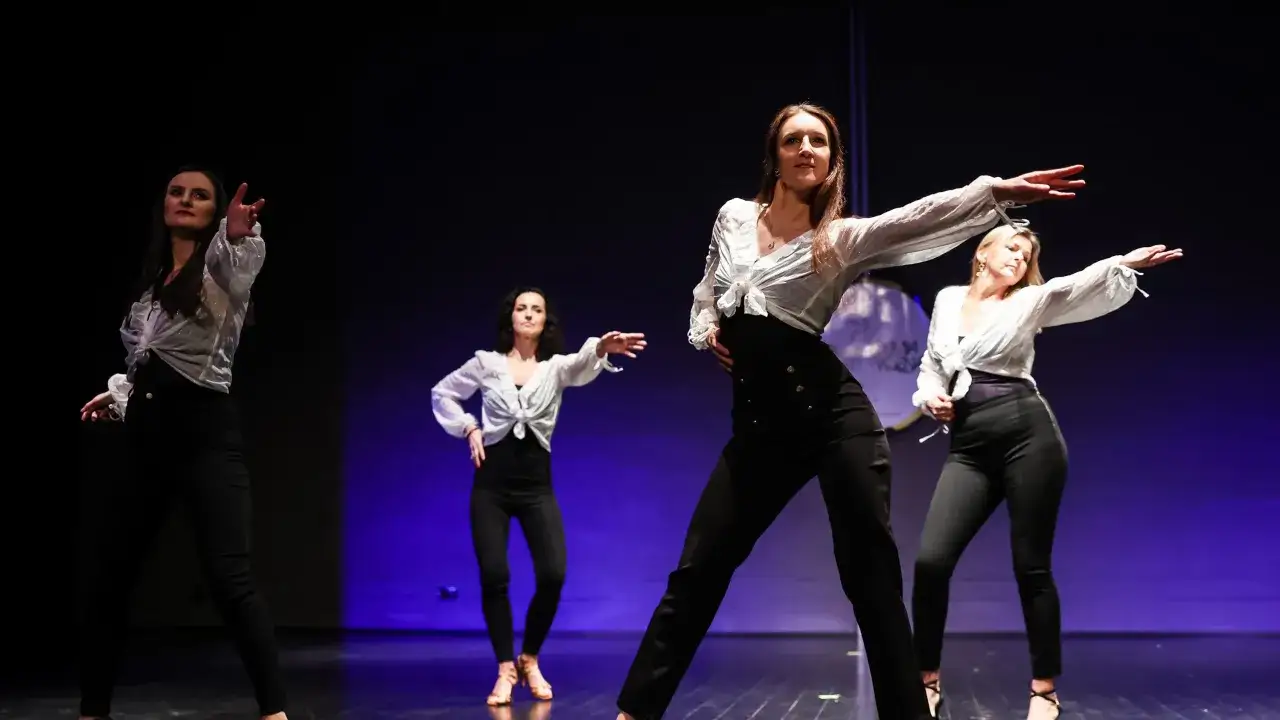 Kobiety w białych koszulach i czarnych spodniach tańczą na scenie. Pokazują, jak tańczyć solo na dyskotece, pewne siebie i pełne gracji.