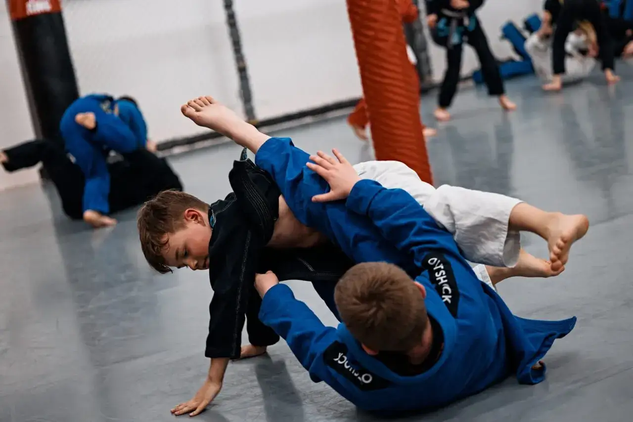 Dzieci ćwiczą jujitsu, walcząc na macie w niebieskich i czarnych kimonach.