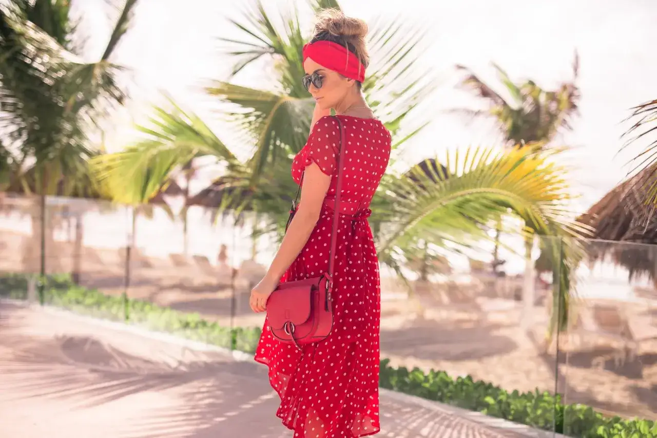 Kobieta w zwiewnej, **czerwonej sukience boho** w białe grochy, z czerwoną opaską na głowie, pozuje na tle palmy i plaży.