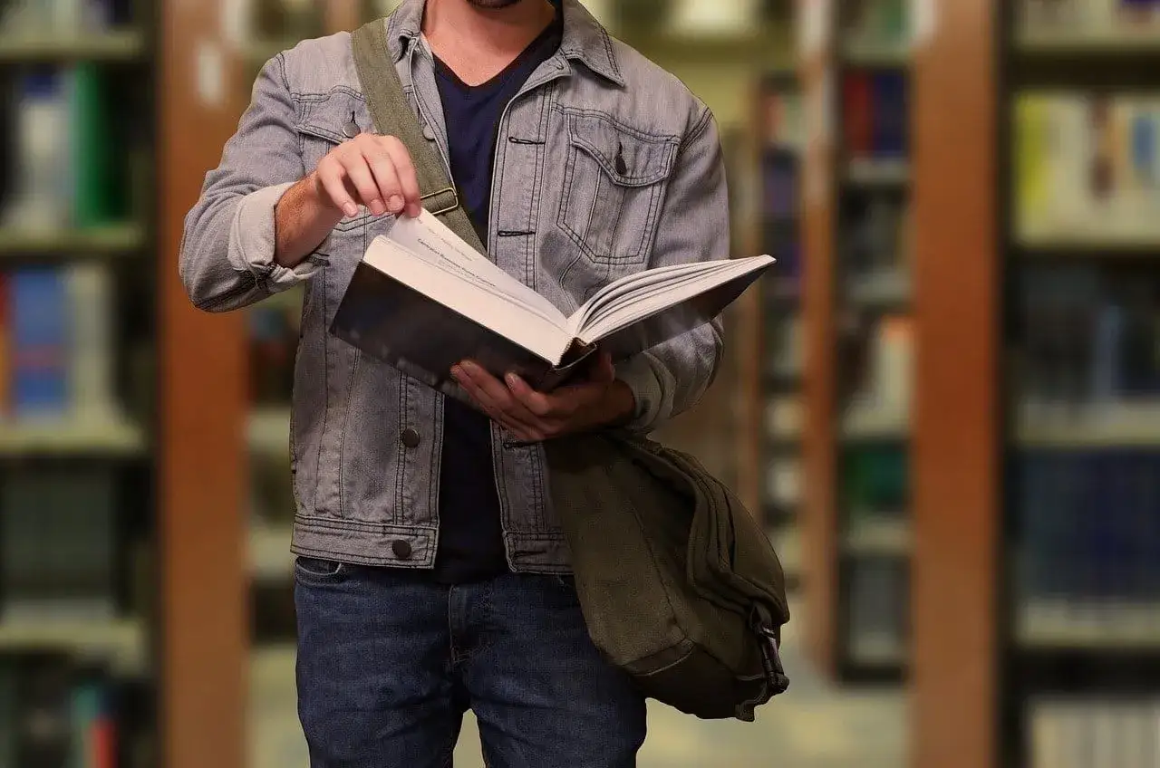 Student w dżinsowej kurtce przegląda książkę w bibliotece. W tle widać regały z książkami.