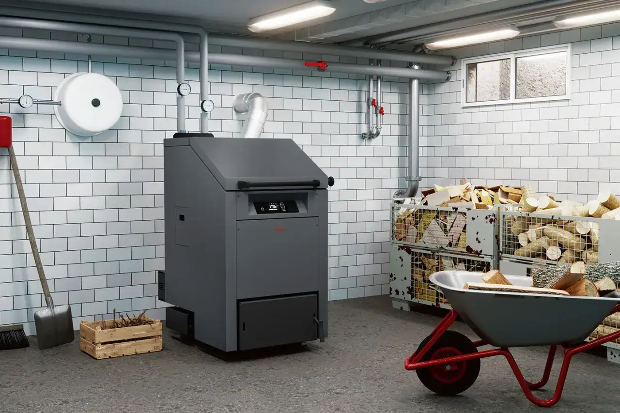 Kotłownia z nowoczesnym kotłem na biomasę, przygotowanym do ogrzewania domu. Drewno w skrzyniach i taczce czeka na załadunek.