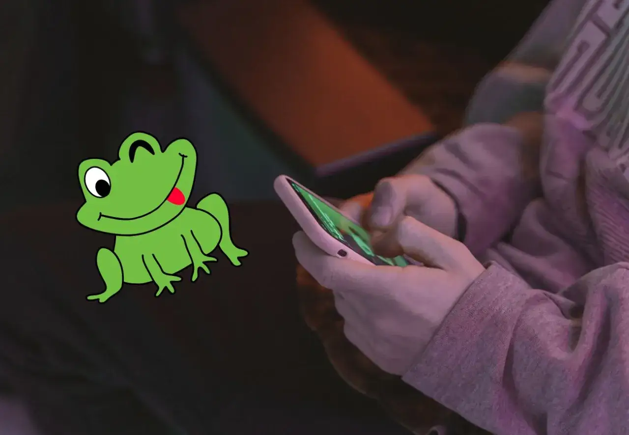 Zielona żaba z wywieszonym językiem obok dłoni trzymających smartfon.