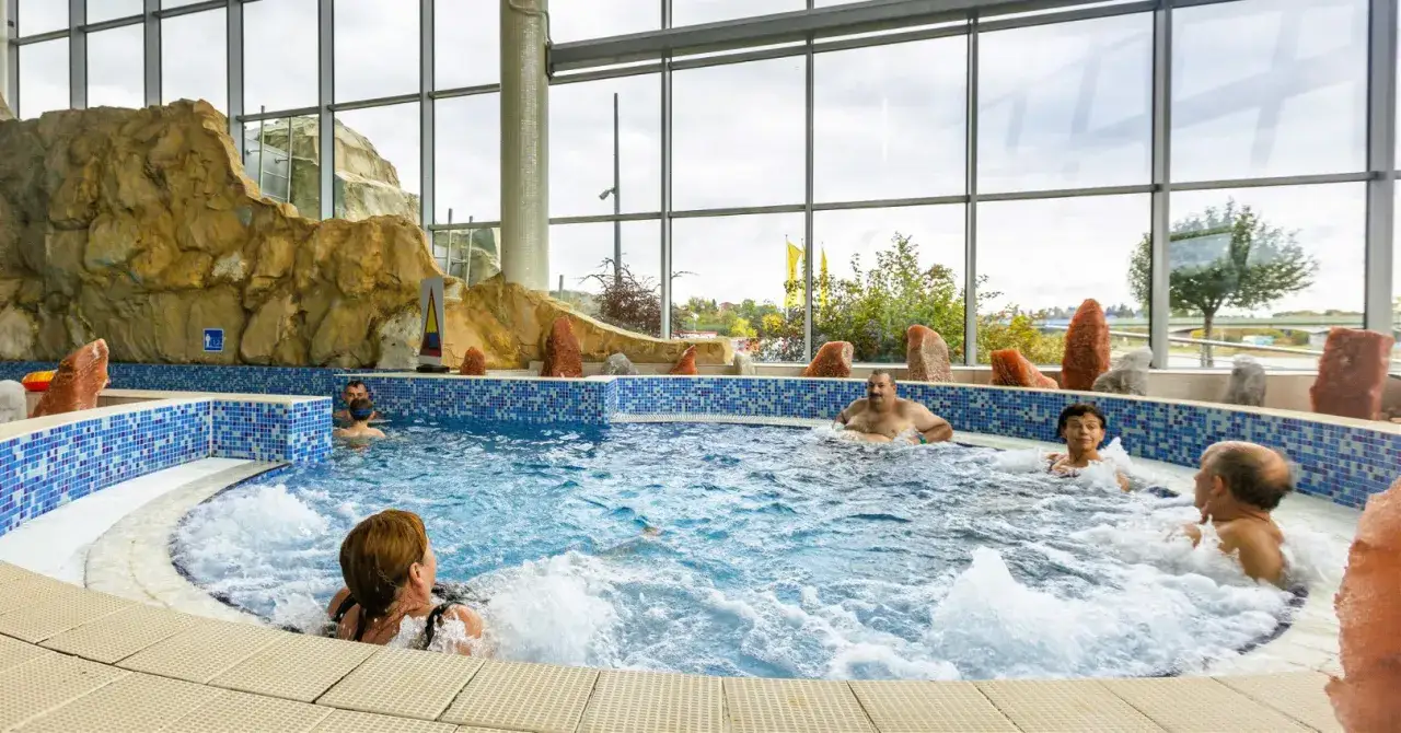 Ludzie relaksują się w basenie termalnym z hydromasażem, otoczonym skalnymi formacjami i dużymi oknami.