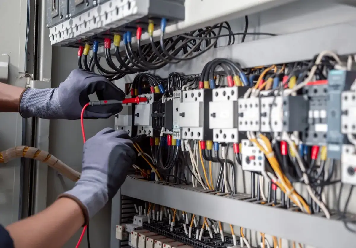 Elektryk w szarych rękawicach mierzy napięcie w skrzynce z przewodami.