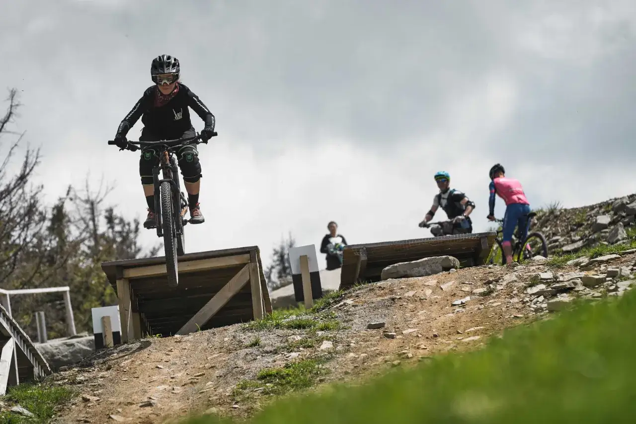 Młoda rowerzystka w kasku i goglach skacze na rowerze w Szczyrk Bike Park. W tle inni rowerzyści.