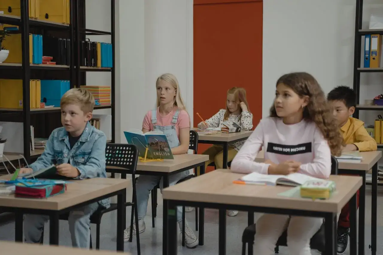 Dzieci siedzą w klasie, skupione na lekcji. Chłopiec w dżinsowej kurtce, dziewczynka z książką po angielsku, a z tyłu inne dzieci. Wszyscy mieli nadzieję, że lekcja szybko się skończy.