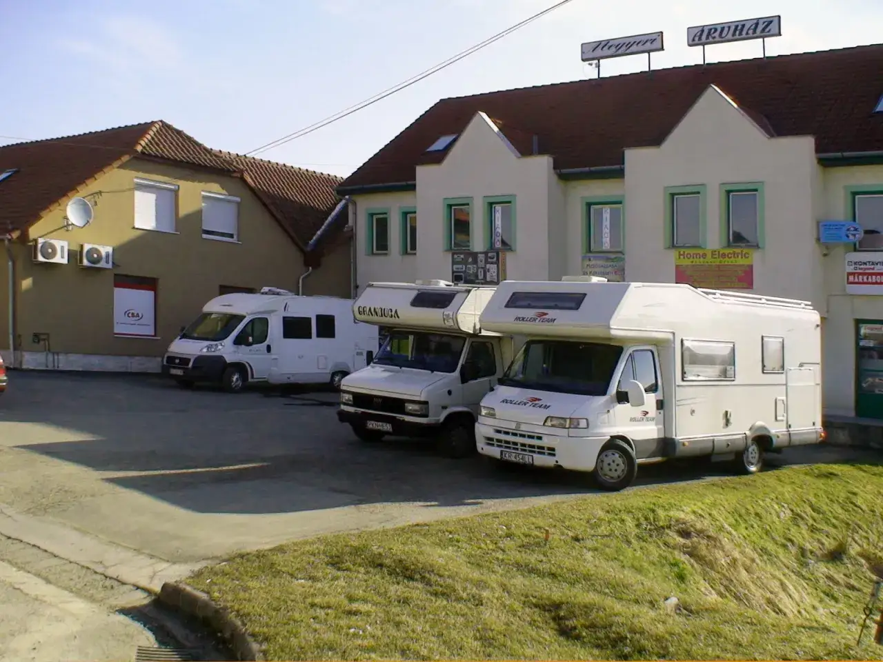 Trzy białe kampery zaparkowane przed budynkami z szyldami.
