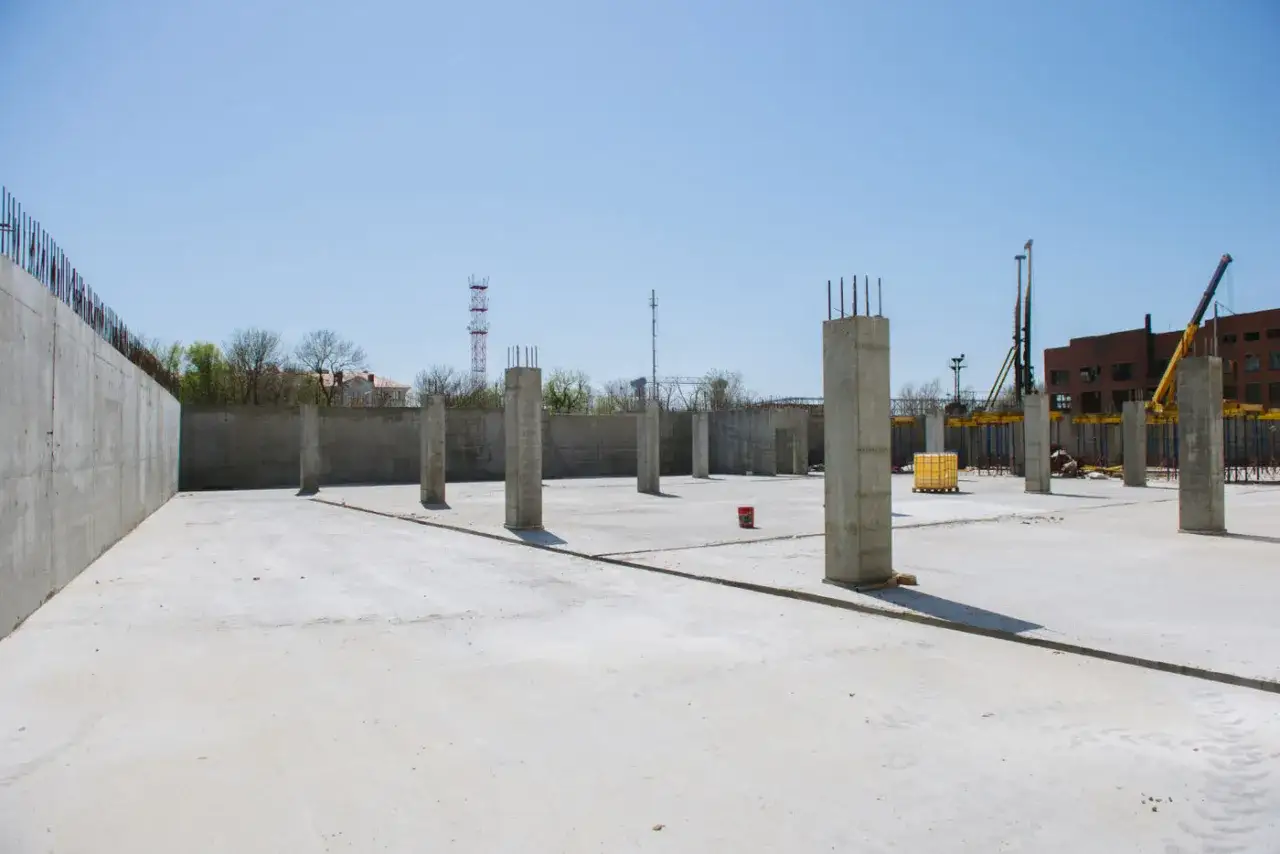 Plac budowy z betonową płytą i słupami betonowymi konstrukcyjnymi. W tle budynek i dźwig.