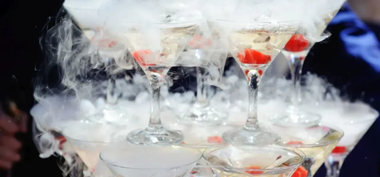 Efektowne drinki z suchym lodem tworzą mglistą, tajemniczą atmosferę. Kieliszki wypełnione są złotym płynem i ozdobione czerwonymi owocami.