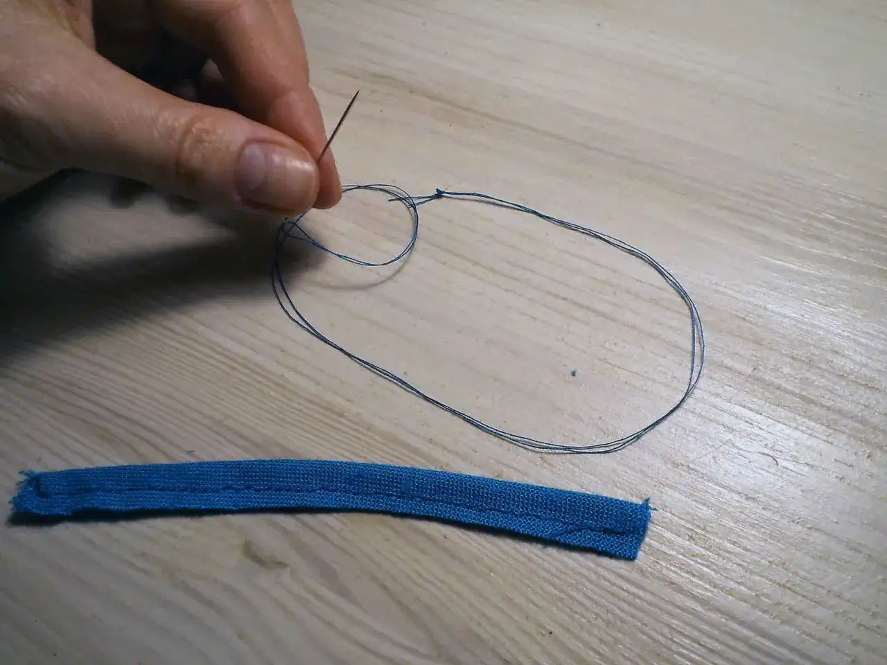 Dłoń trzyma igłę z nitką, tworząc pętelkę na brzegu serwetki. Obok leży niebieski pasek materiału.