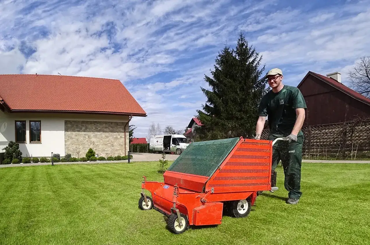 Mężczyzna w zielonym stroju pcha wertykulator, kiedy wertykulujemy trawnik. W tle dom i biała furgonetka.