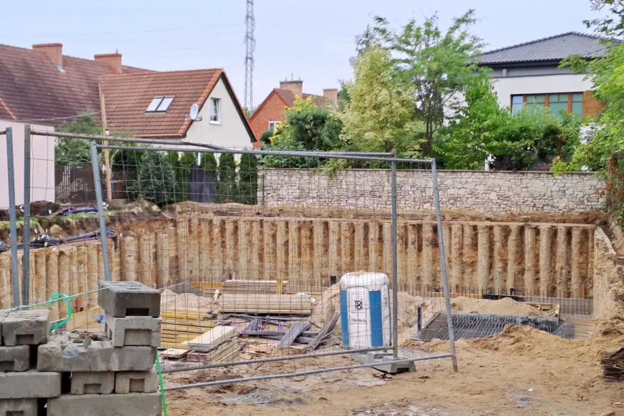 Budowa płyty fundamentowej: widok wykopu z zabezpieczeniem ścian, materiałami budowlanymi i przenośną toaletą. W tle domy i zieleń.