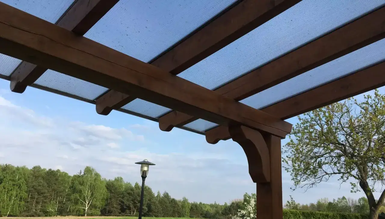 Drewniana pergola z przezroczystym zadaszeniem z poliwęglanu na tle nieba i drzew.