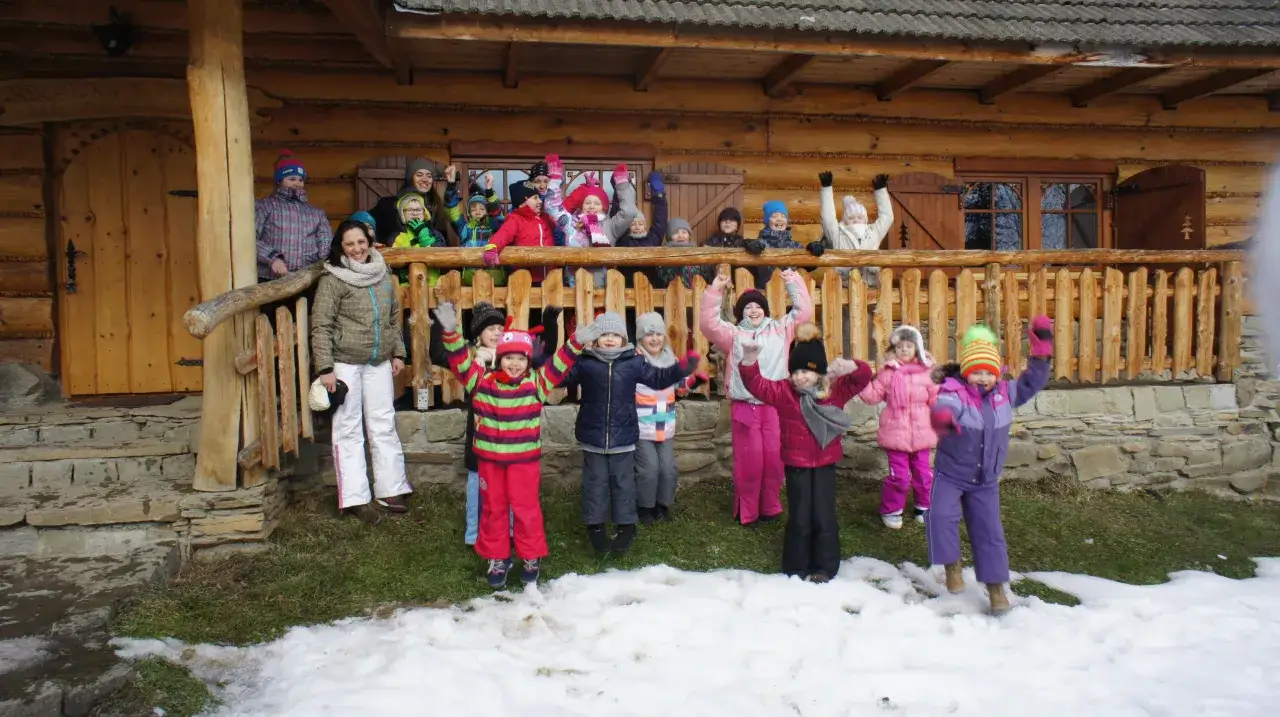 Wesoła gromadka dzieci pozdrawia z bacówki u harnasia. Wszyscy ubrani w zimowe stroje, radośnie machają rękami na tle drewnianego domu.
