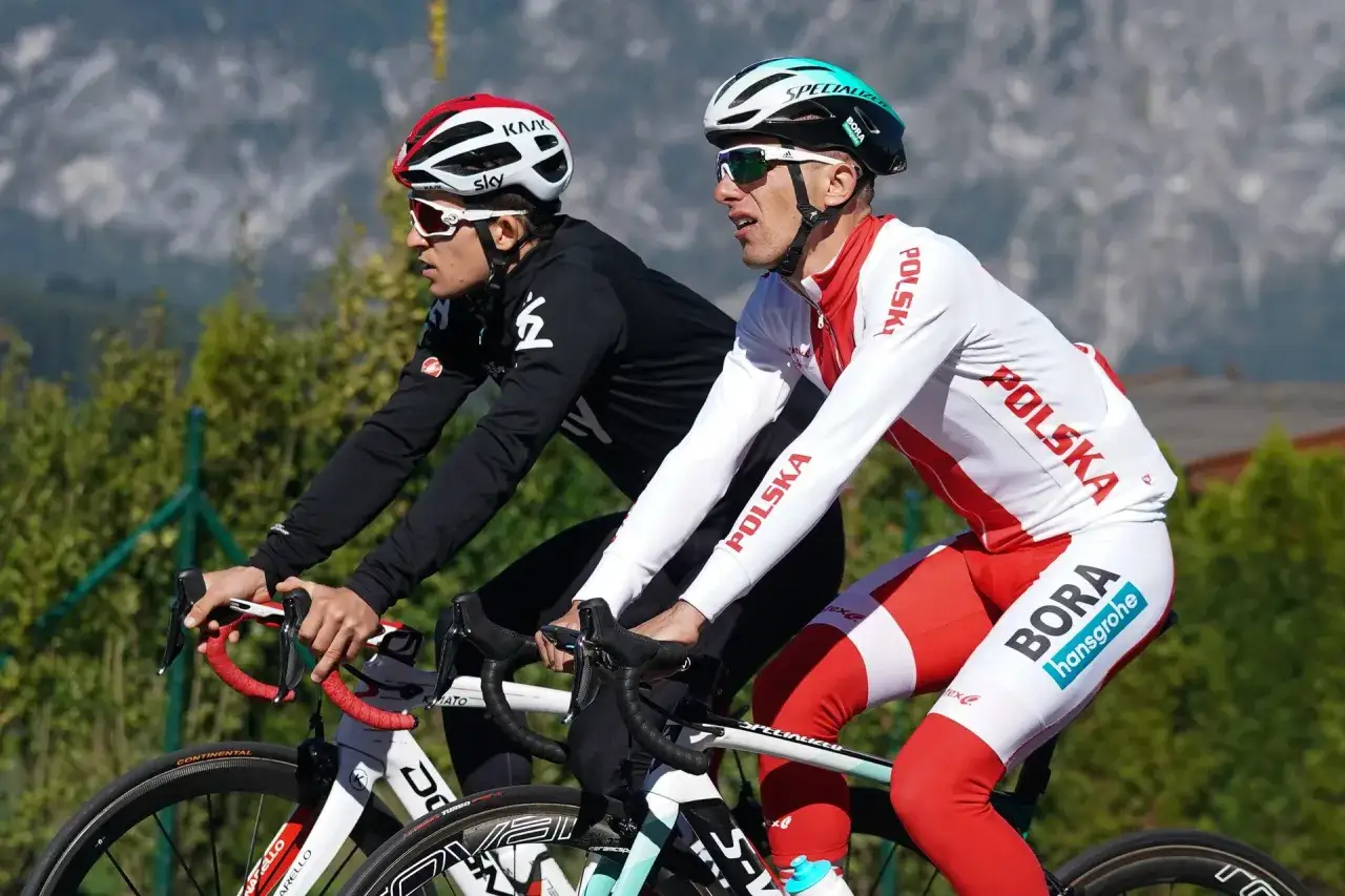 Dwóch kolarzy, być może najlepsi kolarze w historii, w biało-czerwonych strojach z napisem "Polska" i logiem "Bora Hansgrohe", ściga się na tle gór.