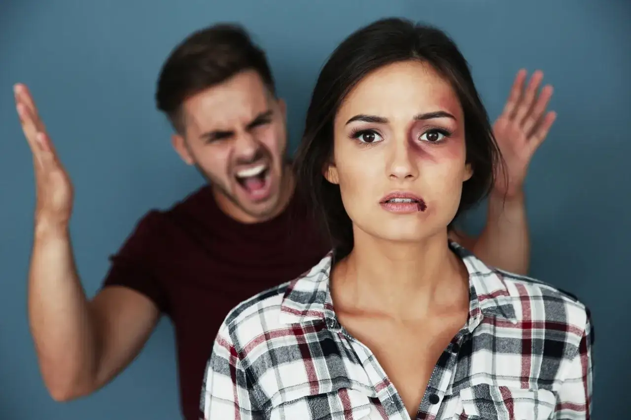 Kobieta z siniakami na twarzy i rozdartymi ustami, za nią krzyczący mężczyzna.