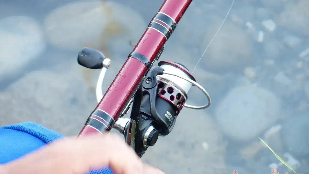 Wędka z kołowrotkiem czeka na lipcowe brania. Woda i kamienie w tle.