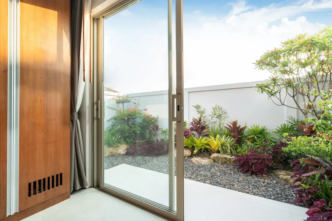 Uchylno przesuwne drzwi tarasowe otwierają widok na bujny, tropikalny ogród z kolorowymi roślinami i kamieniami.