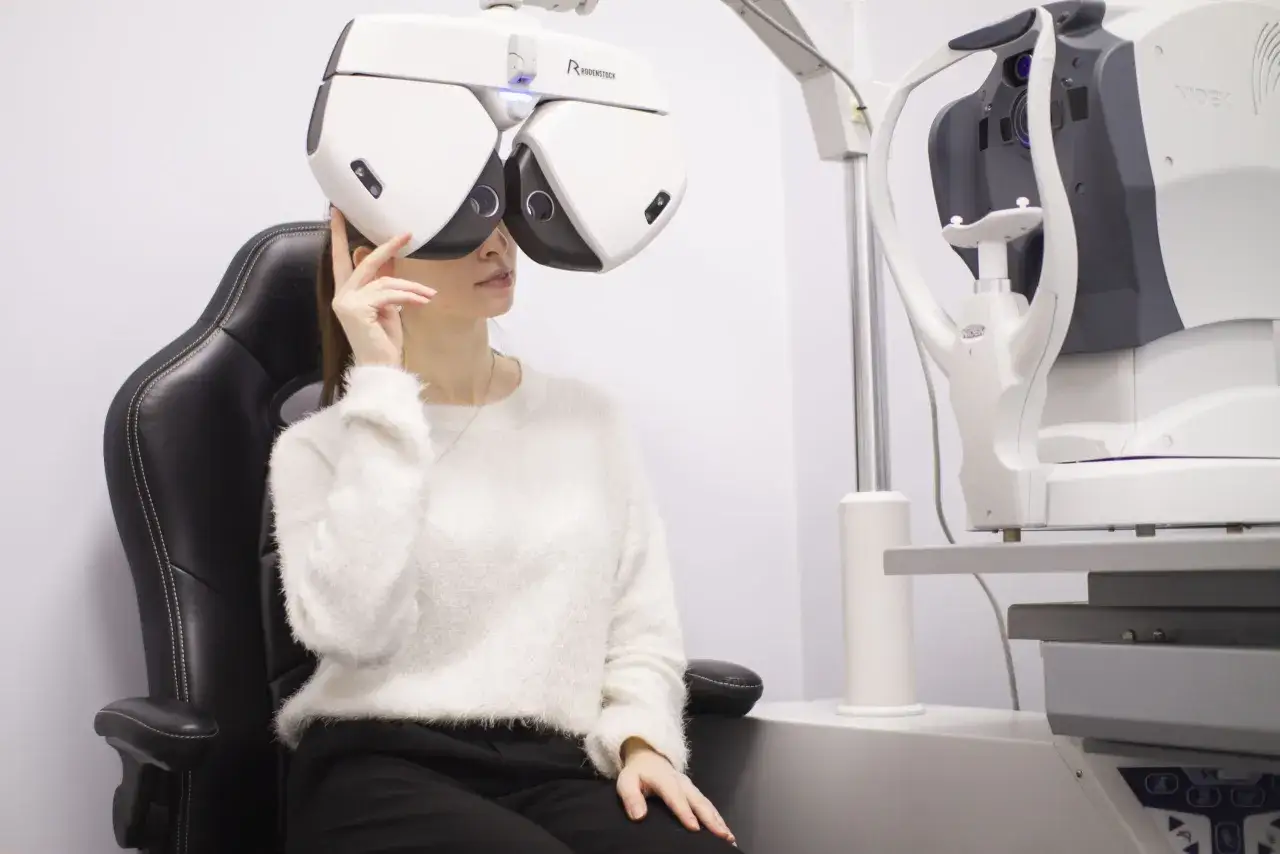 Kobieta w białym swetrze siedzi na czarnym fotelu, badana przez optometrystę na nowoczesnym sprzęcie.