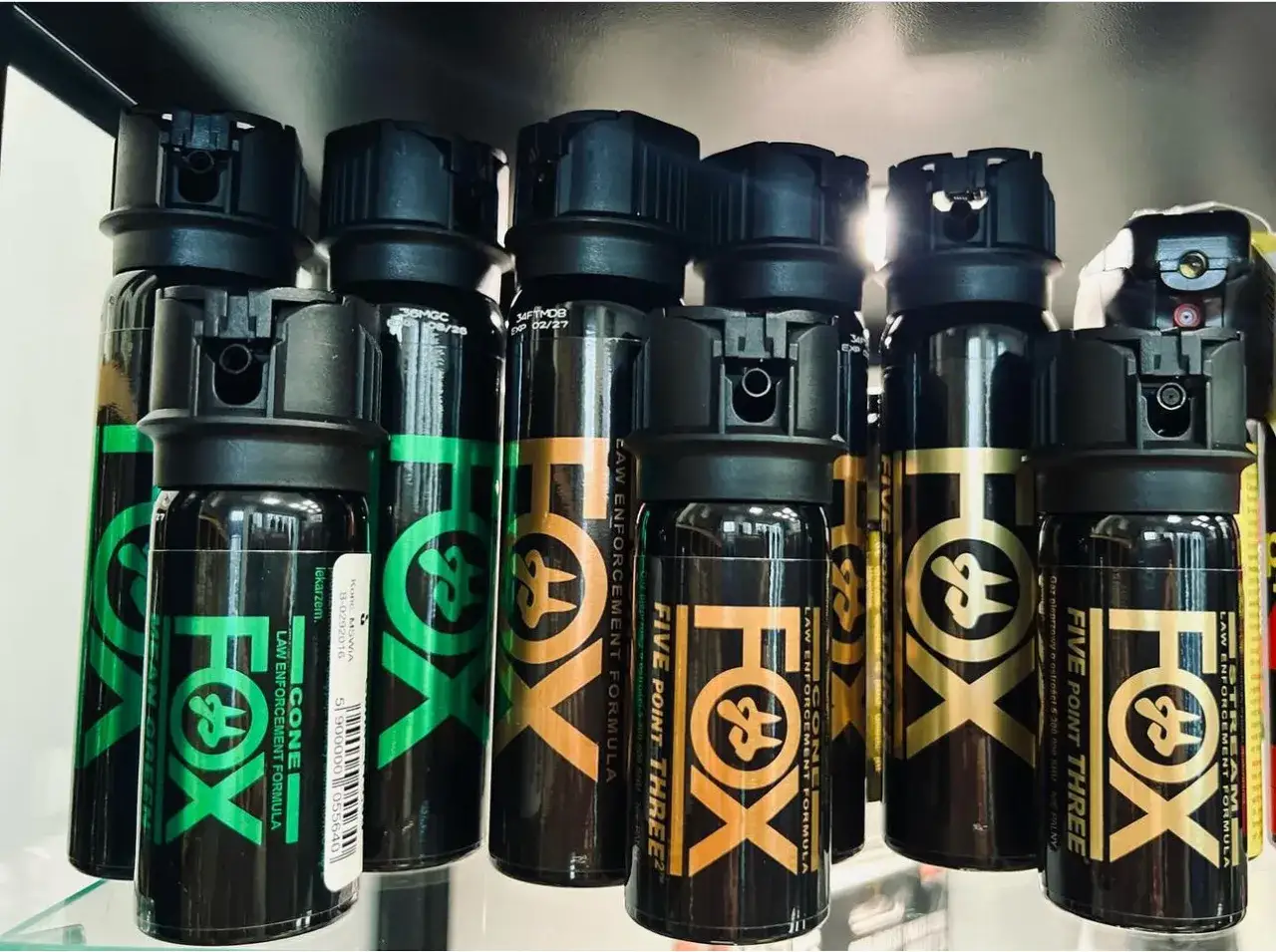 Kilka puszek gazu pieprzowego marki FOX z czarnymi nakrętkami, niektóre z zielonymi, inne z pomarańczowymi lub złotymi napisami.