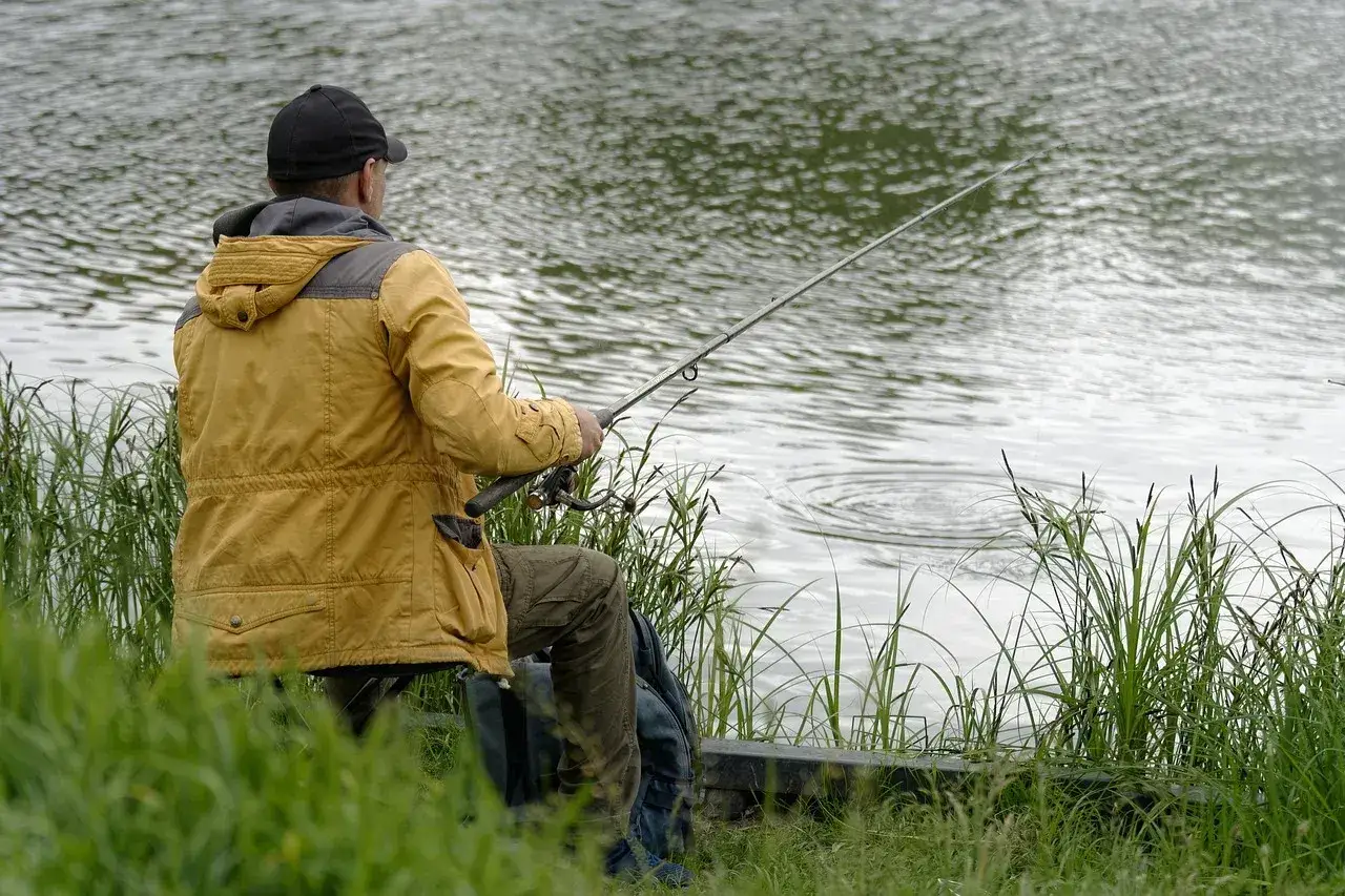 Wędkarz nad jeziorem, przygotowuje zanętę na ryby. Woda faluje, a trawa otacza brzeg.