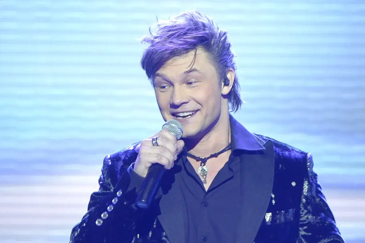 Jaka to melodia piosenkarze: przystojny mężczyzna z fioletowymi włosami śpiewa do mikrofonu w błyszczącej marynarce.