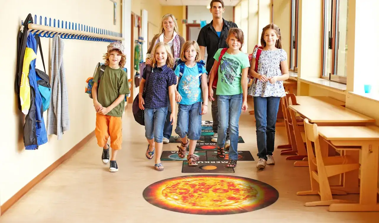 Dzieci i dorośli idą szkolnym korytarzem, stąpając po planszy z planetami. Fajne inicjatywy w szkole promują naukę przez zabawę.