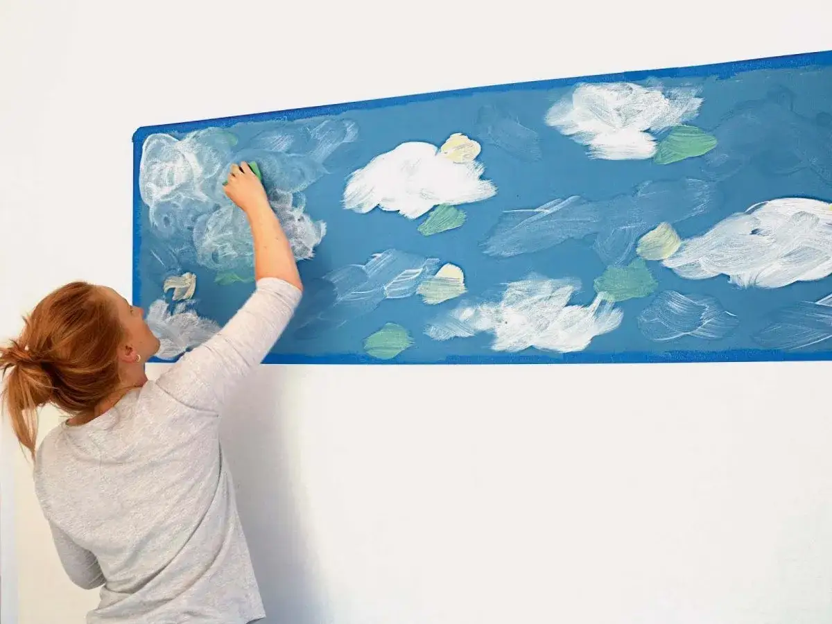 Kobieta maluje gąbką białe chmury na niebieskiej ścianie. Prosty sposób, jak namalować chmury na ścianie, tworząc niebo.