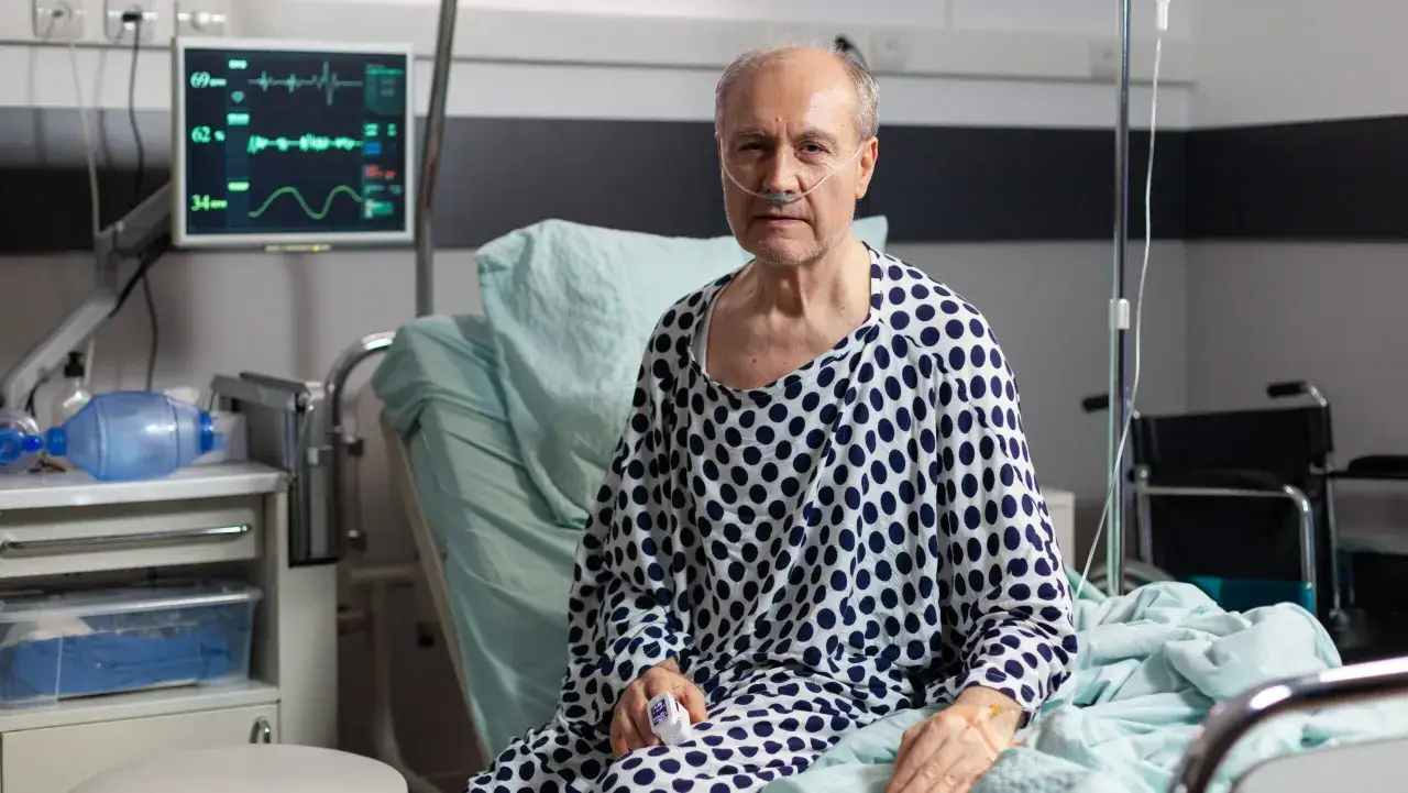 Starszy mężczyzna w szpitalnym łóżku, z tlenem w nosie, walczy z ostrą białaczką szpikową. Monitor pokazuje jego parametry życiowe.