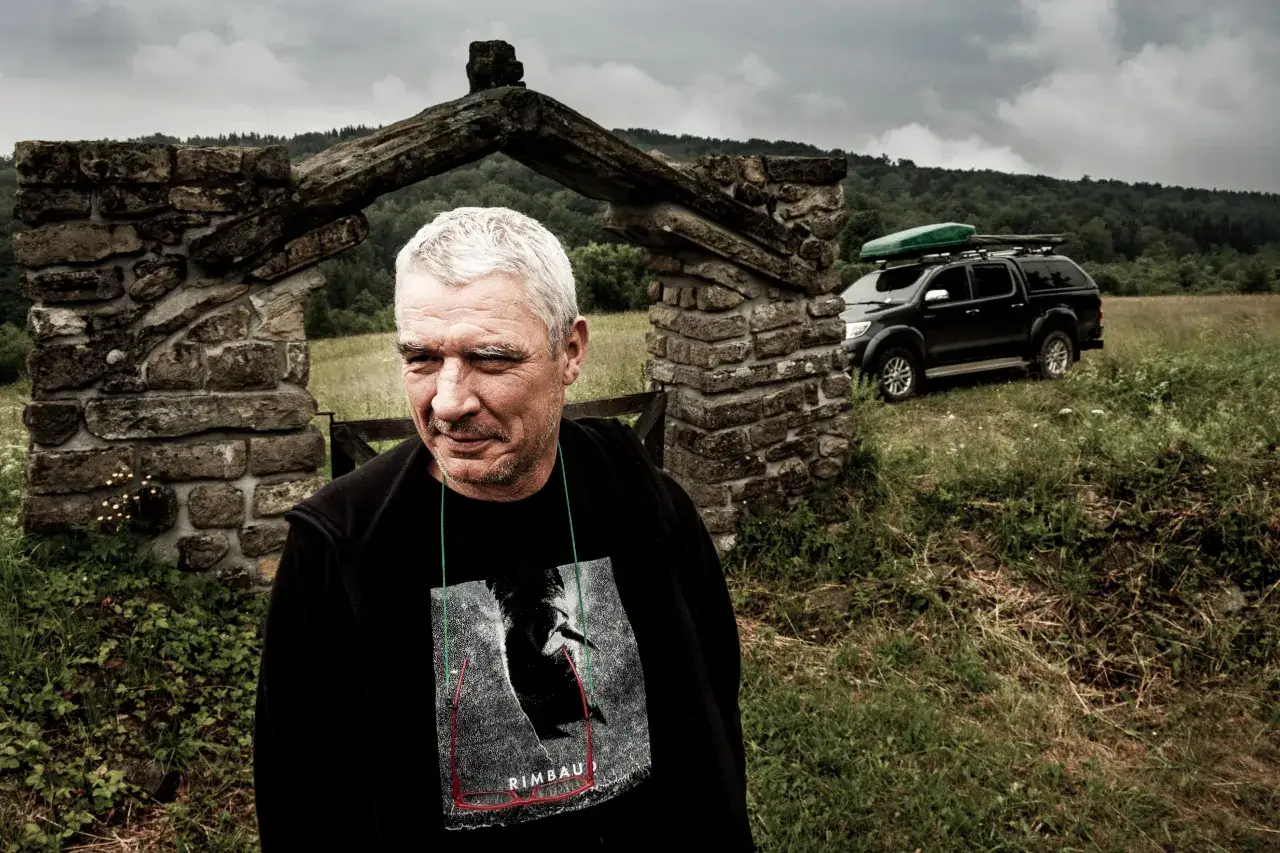 Mężczyzna z siwymi włosami przed kamiennym łukiem, za nim czarny SUV z bagażnikiem dachowym.