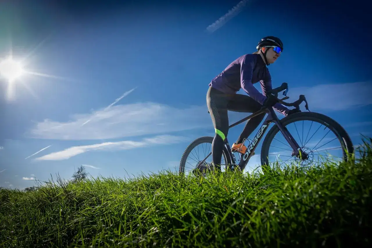 Kobieta w kasku i okularach przeciwsłonecznych odbywa trening na rowerze gravelowym, pokonując pagórkowaty teren porośnięty zieloną trawą.