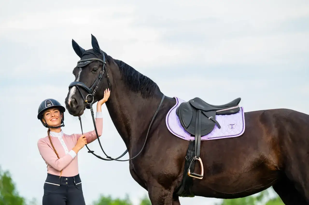 Uśmiechnięta jeźdźczyni w kasku i bluzce w kolorze pudrowego różu stoi obok ciemnobrązowego konia z fioletową czaprakiem.