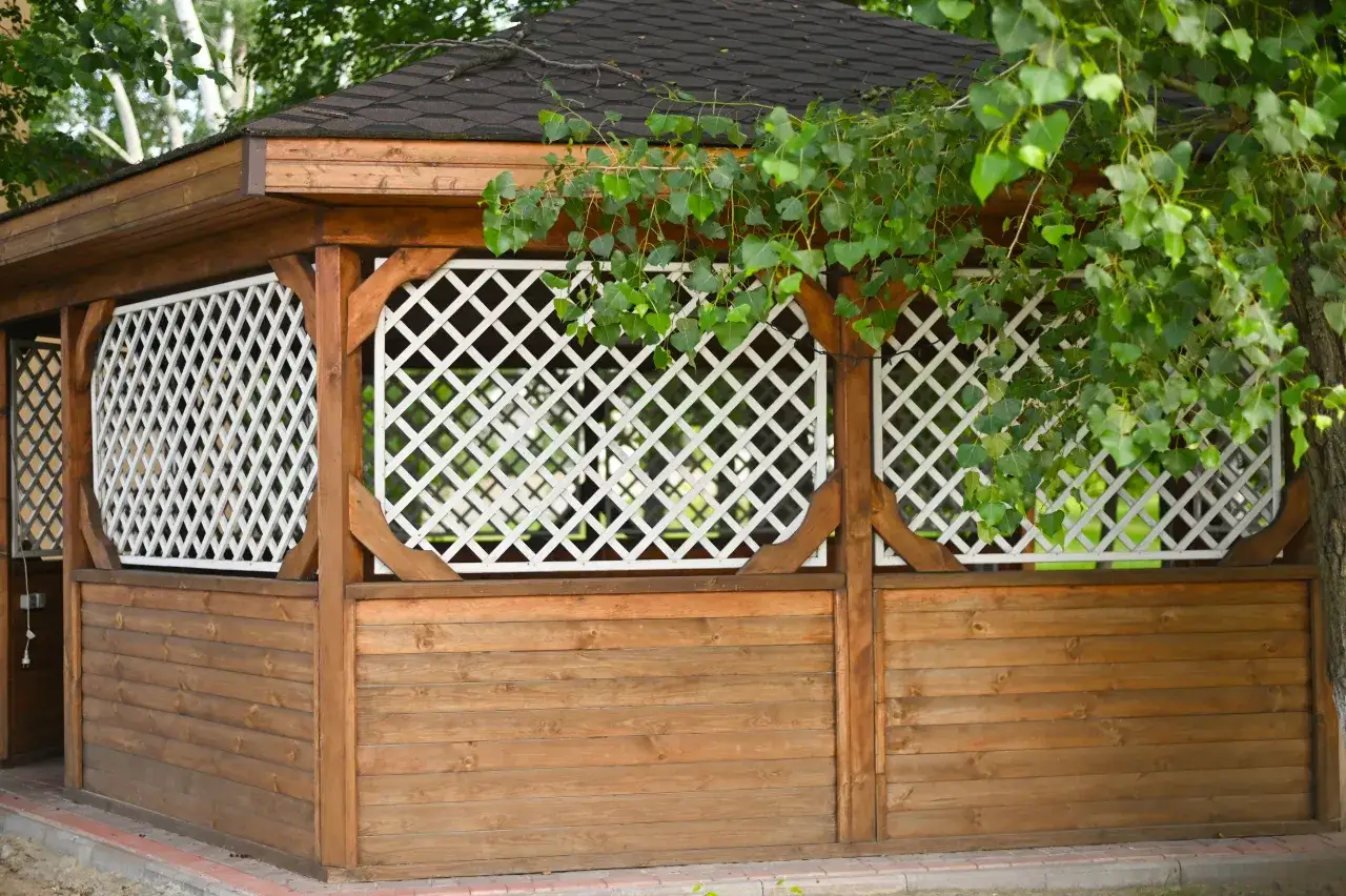 Drewniana altana z ażurowymi panelami i zielonymi liśćmi drzew.