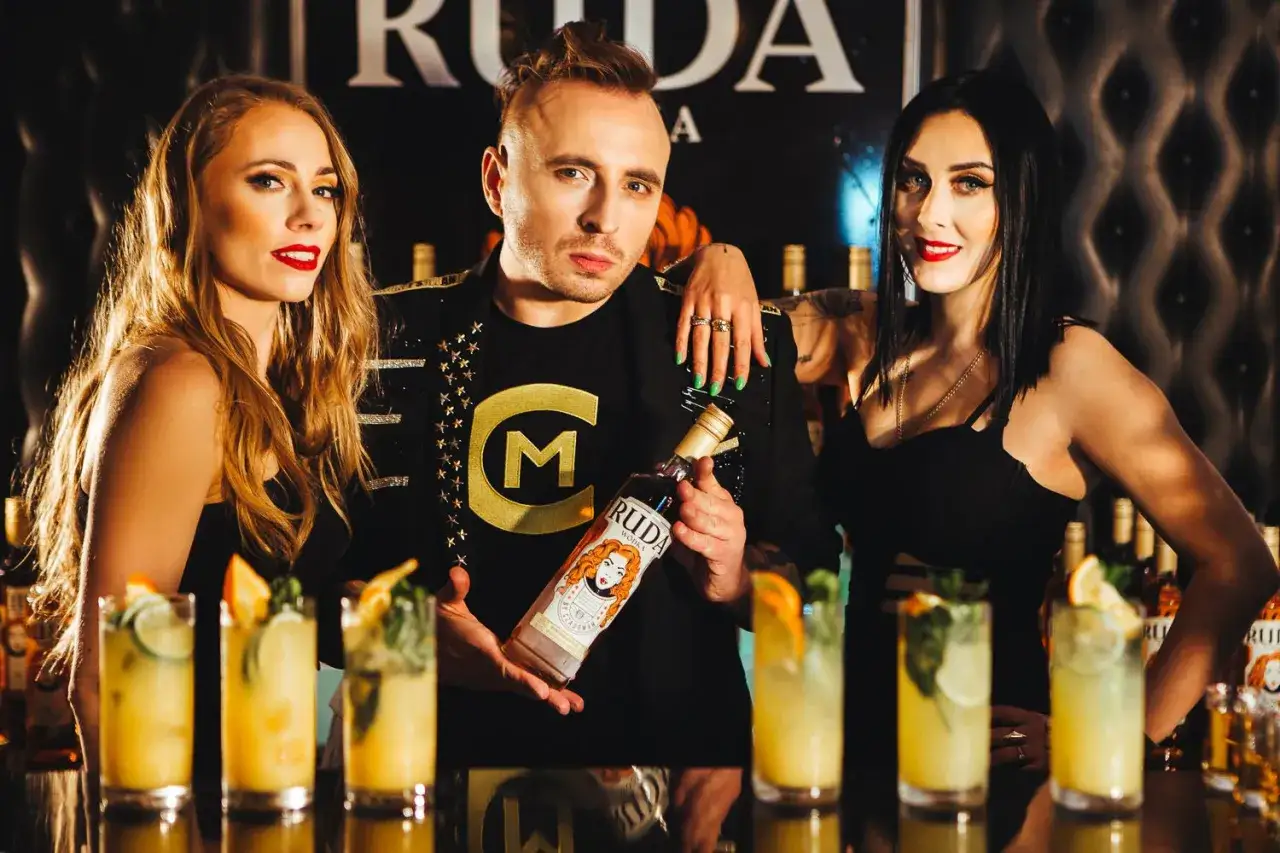 Trzy osoby pozują z butelką wódki Ruda. W tle widać drinki i logo.