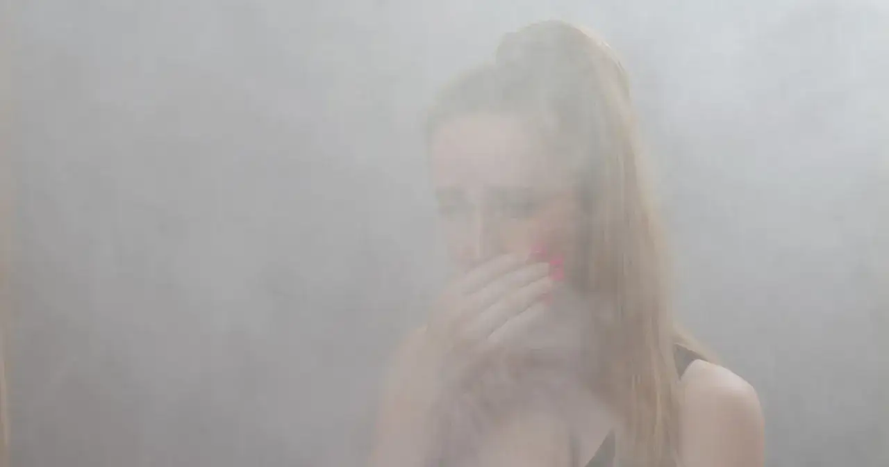 Kobieta zakrywa usta dłonią, otoczona gęstym dymem. Czy można się zatruć dymem z kominka? To pytanie nabiera tu realnego znaczenia.