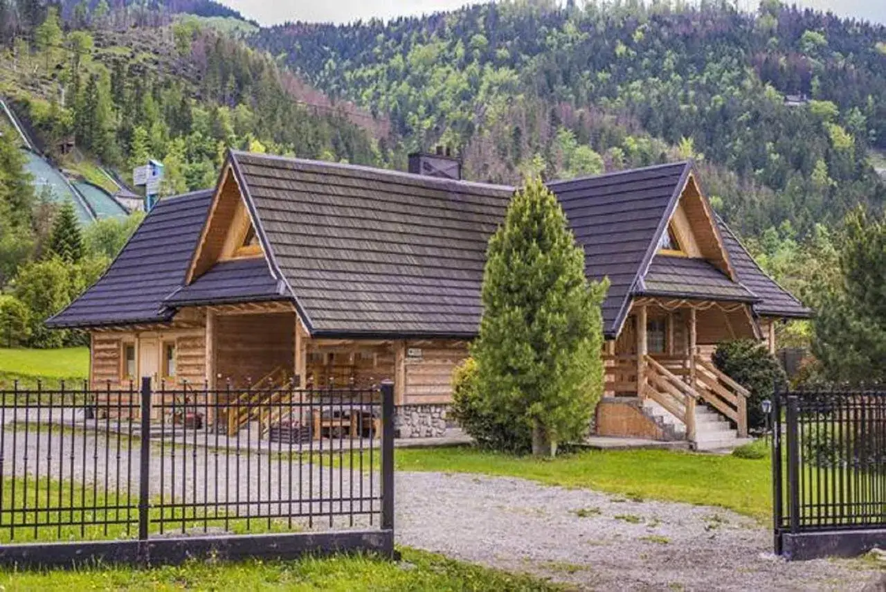 Drewniana bacówka pod skocznią, otoczona zielenią, z widokiem na górskie zbocza.