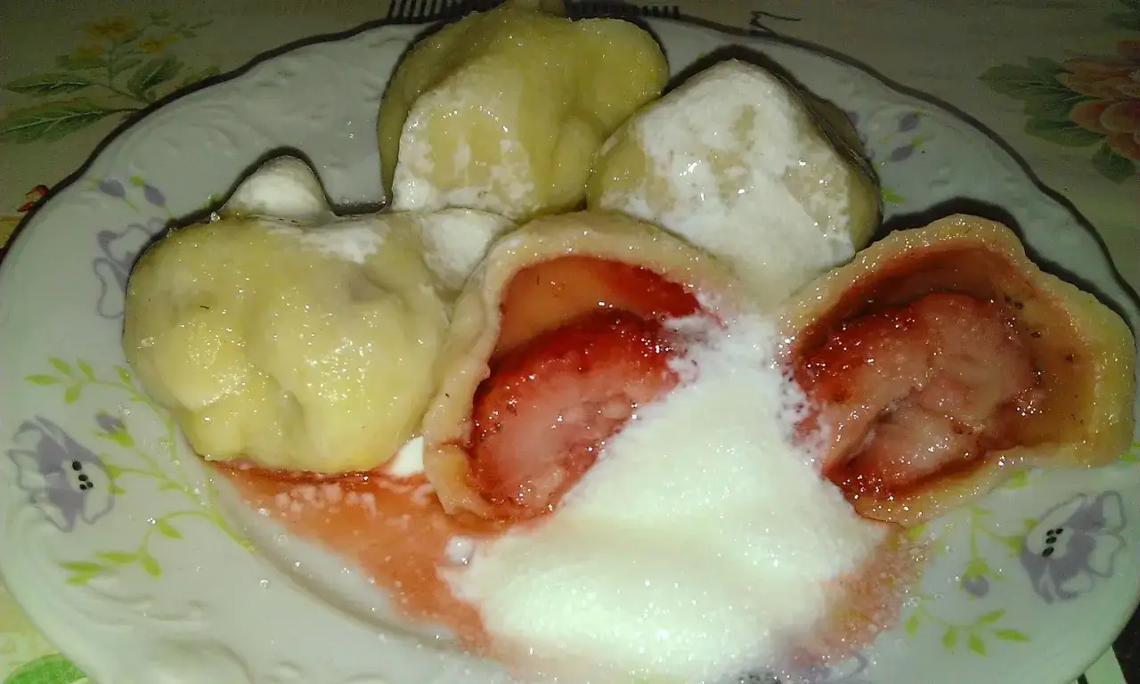 Jak gotować pierogi mrożone z truskawkami? Na talerzu pyszne, gorące pierogi z owocami, polane śmietaną i cukrem.