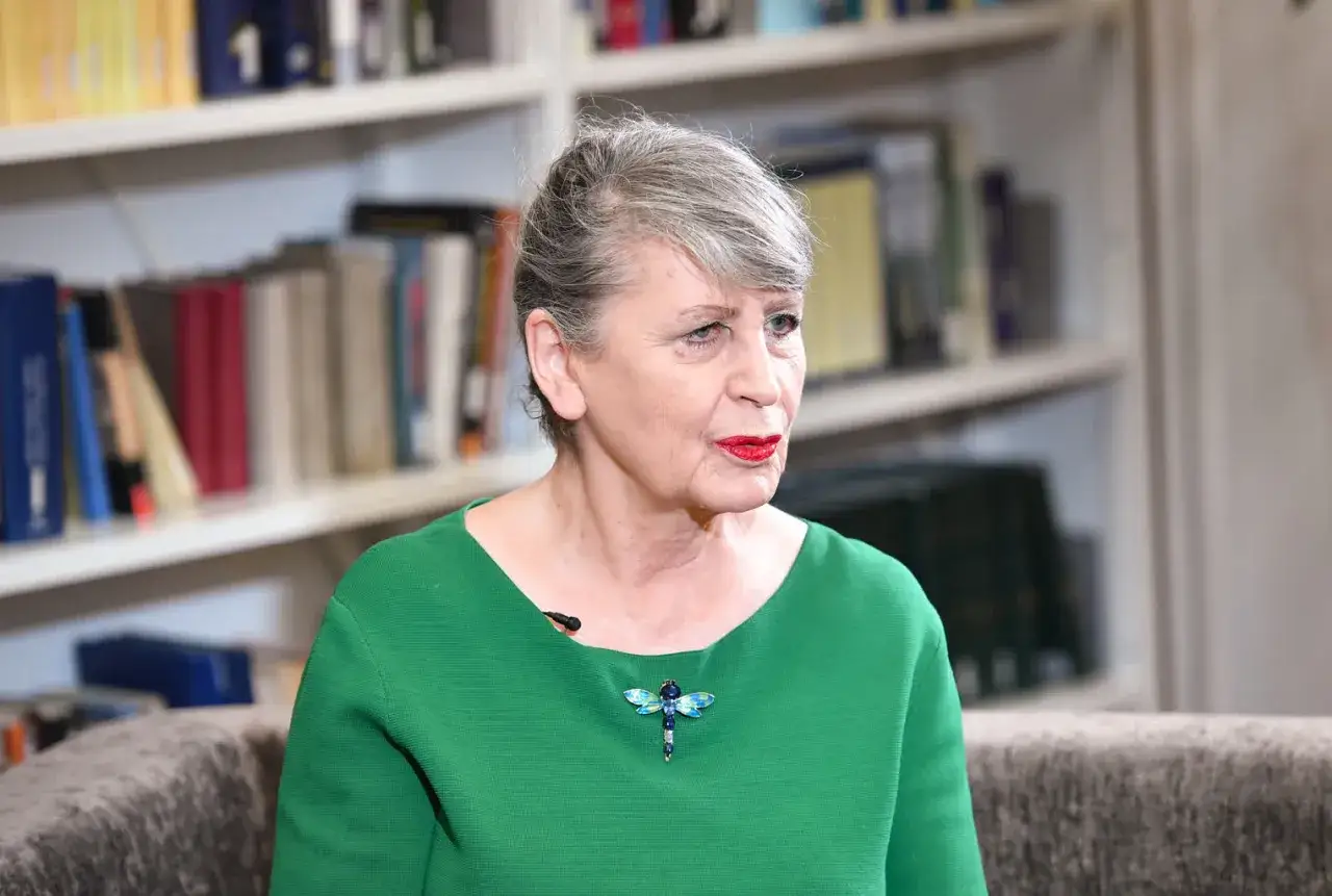 Joanna Wnuk-Nazarowa w zielonej bluzce z broszką w kształcie ważki, na tle regałów z książkami.