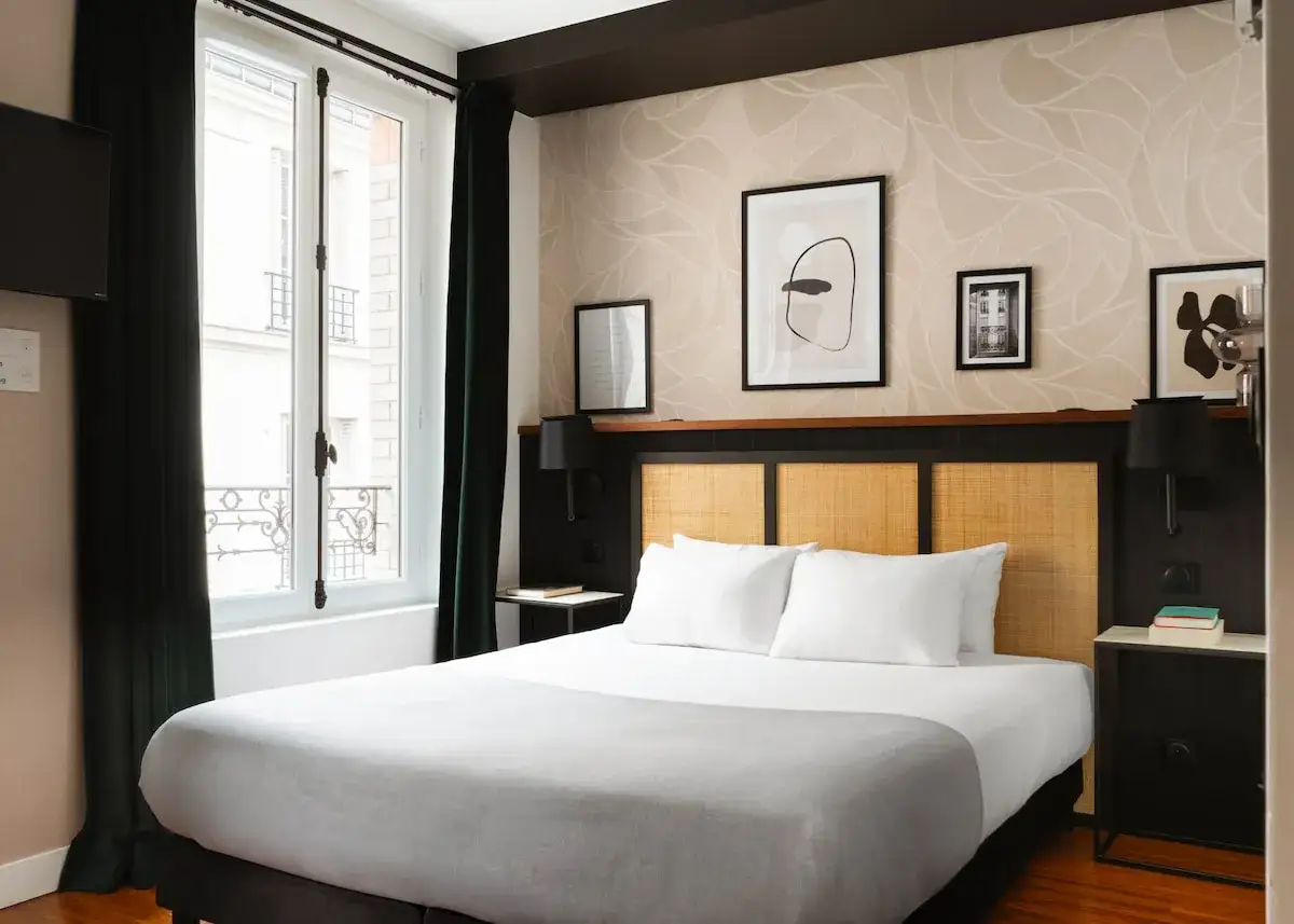 Przytulna sypialnia w 17 dzielnicy Paryża. Łóżko z białymi poduszkami i szarym kocem, okno z widokiem na paryskie kamienice.