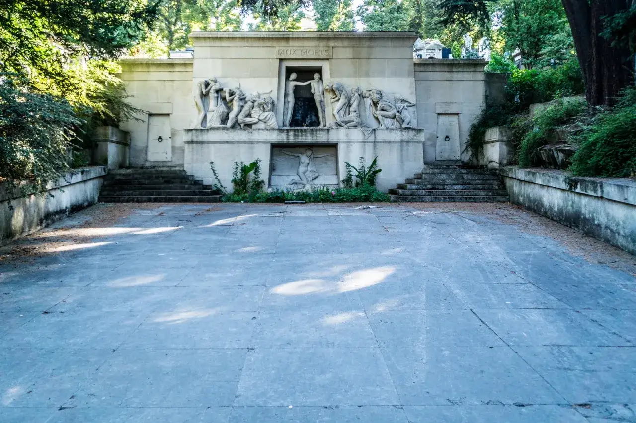 Monumentalna fasada z płaskorzeźbami na cmentarzu Père Lachaise. Kamienne schody prowadzą do wejścia, a wokół rosną drzewa.
