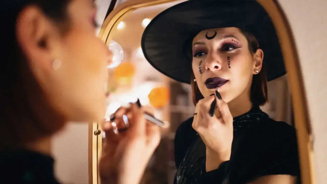 Frau mit Hexenhut und Halloween-Make-up trägt Lippenstift vor einem Spiegel auf.