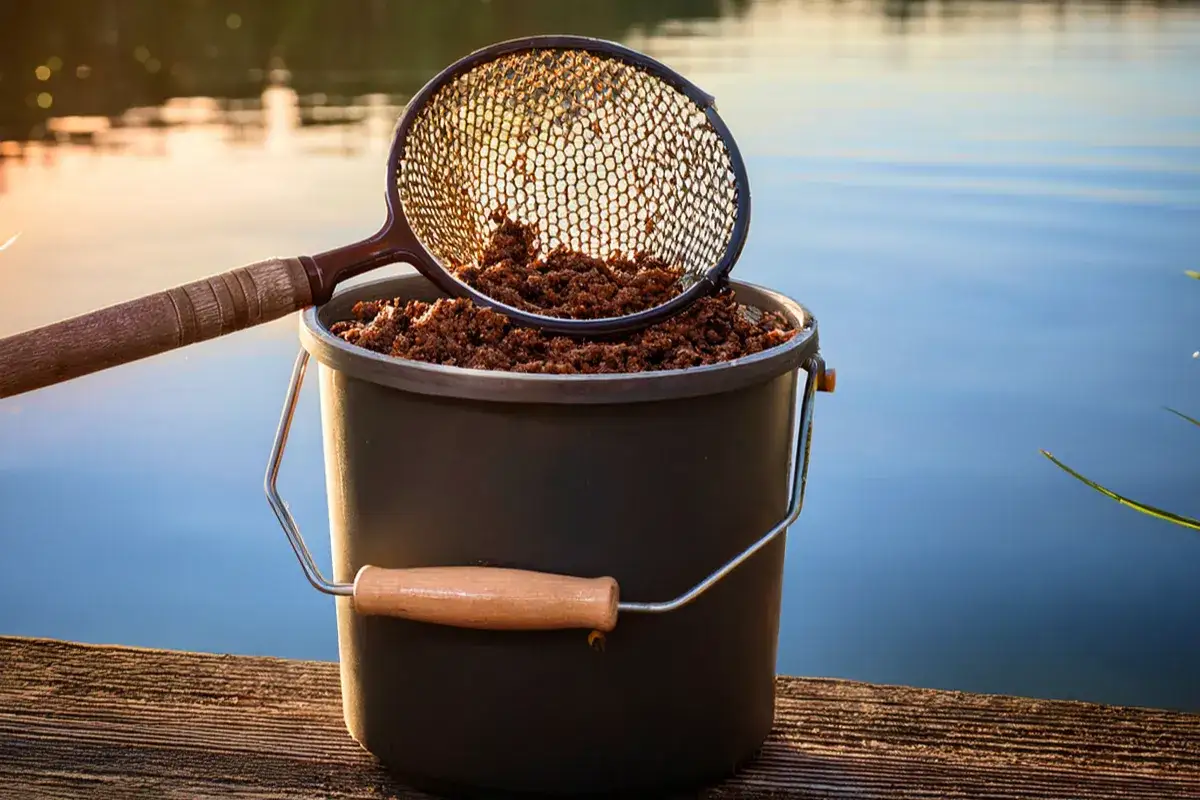 Sito do przecierania zanęty nad brzegiem jeziora. W wiaderku gotowa mieszanka, a sitko właśnie ją odsiewa.