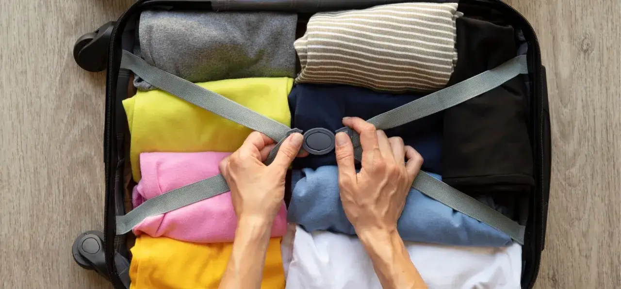 Ręce zapinają pasy w otwartej walizce wypełnionej kolorowymi, złożonymi ubraniami.