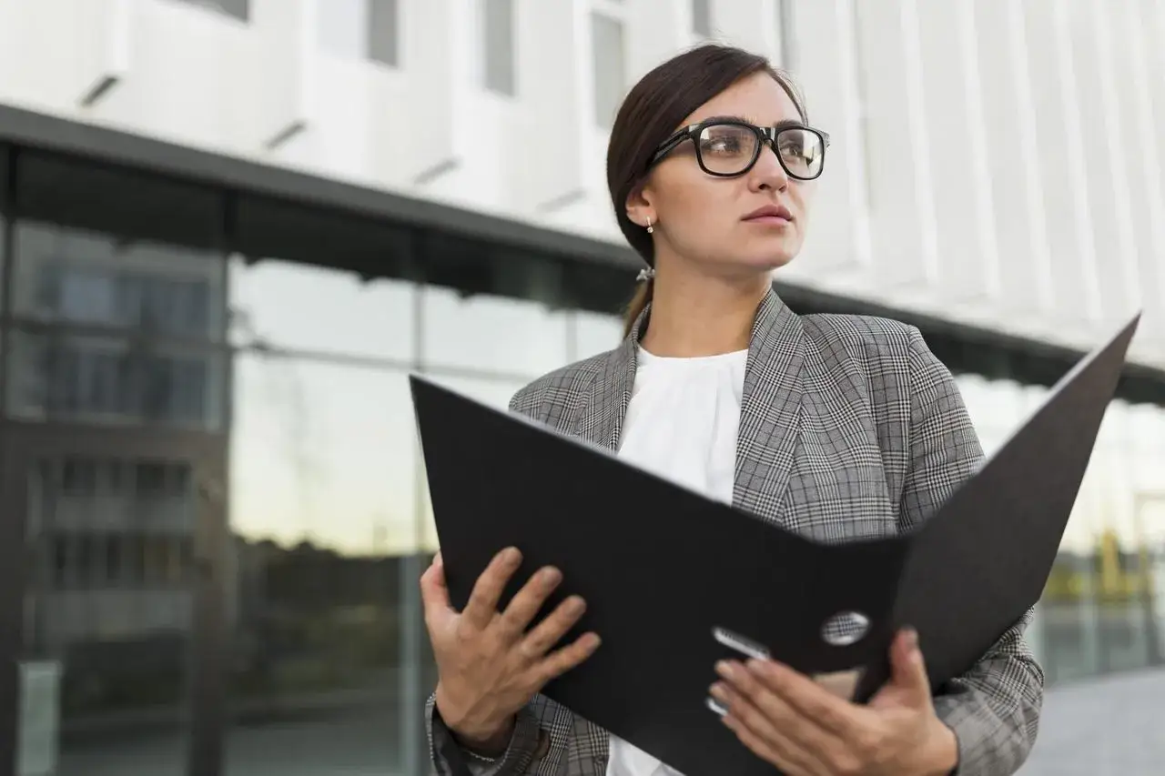 Kobieta w okularach trzyma czarną teczkę przed nowoczesnym budynkiem.