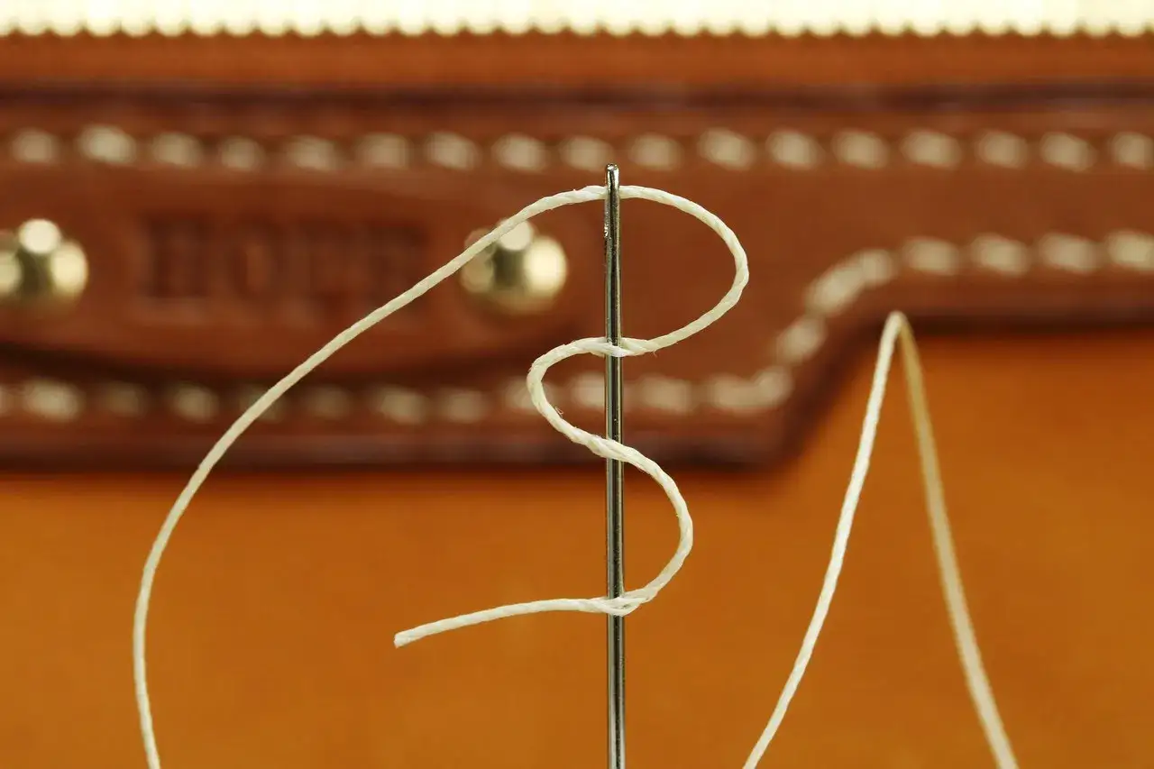 Zbliżenie na igłę do szycia ręcznego z nawleczoną białą nicią, na tle brązowej skóry z ozdobnymi przeszyciami. Różne rodzaje igieł do szycia ręcznego mogą mieć różne zastosowania.